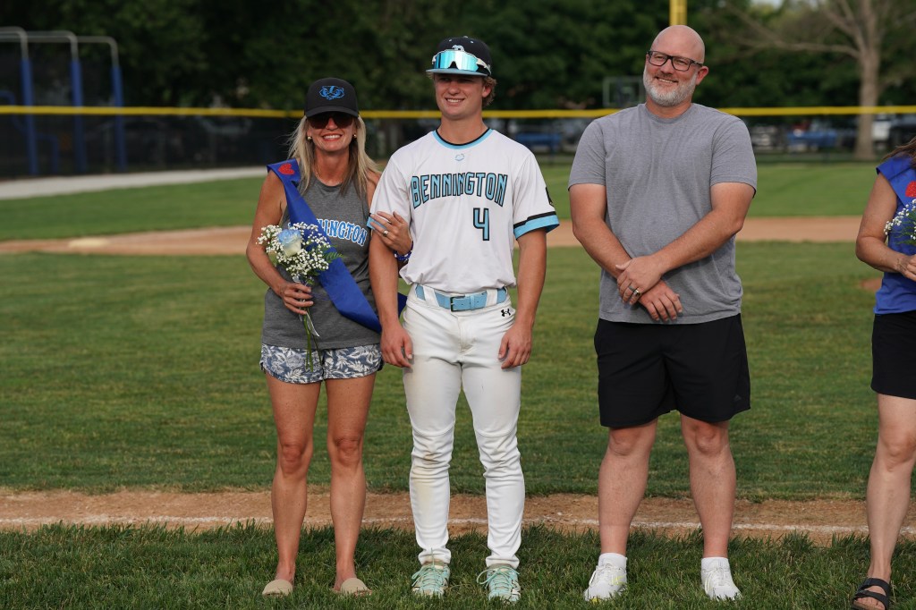 A young male baseball player wearing a team jersey stands on a baseball field alongside two adults holding flowers and wearing sashes, while another adult stands next to them. The setting is a sunny day with a baseball diamond in the background.