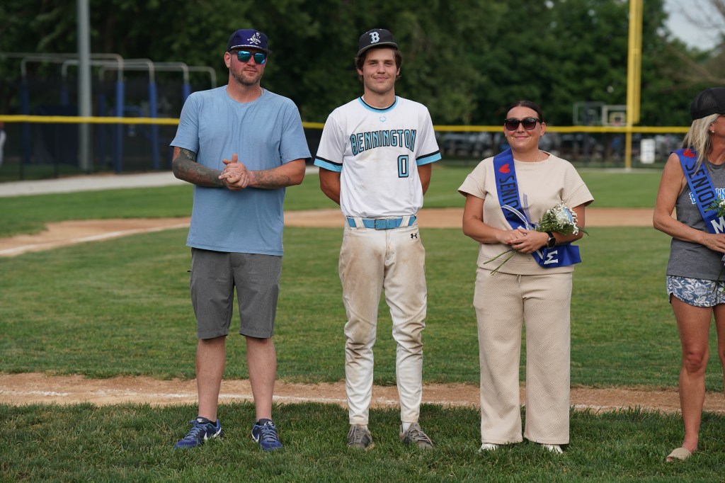 A group of three individuals standing on a baseball field. The first person is a man wearing sunglasses and a blue t-shirt with tattoos on his arms. The second is a young man in a baseball uniform with the number 0, and the third is a woman in a light-colored outfit holding flowers, wearing sunglasses, and a sash that reads 'Senior'.