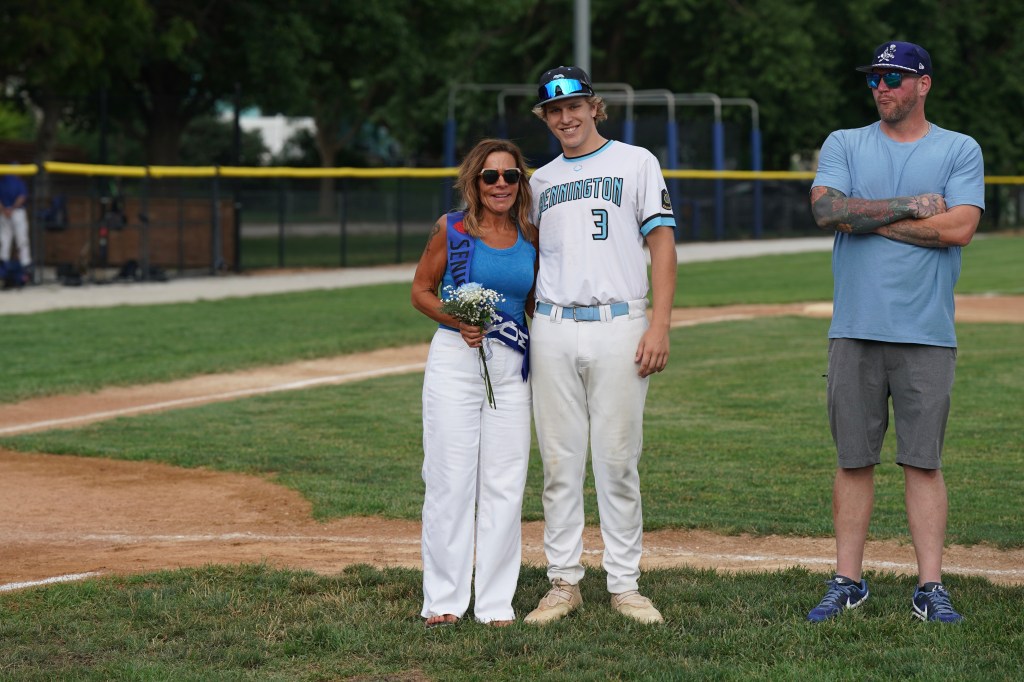 A woman in white pants and a blue top, holding flowers, stands next to a young man in a baseball uniform with the number 3. Both are smiling on a baseball field, with a man in a blue shirt standing nearby.