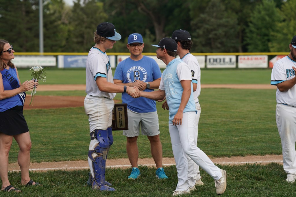 A baseball player in catching gear shakes hands with a teammate while being presented with an award, surrounded by coaches, teammates, and a woman holding flowers.