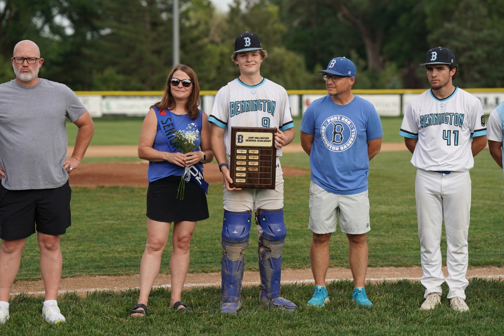 A group of individuals at a baseball field, including a player in a catcher's gear holding a plaque, accompanied by family and coaches.