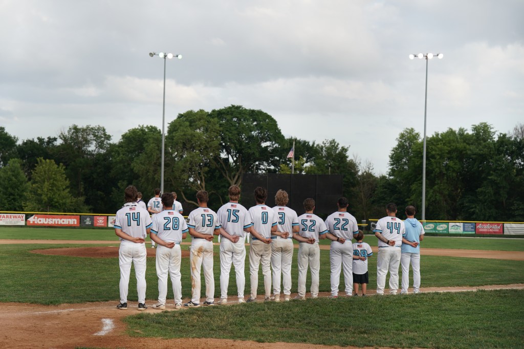 A group of baseball players standing in a line on the field, facing the outfield, with their hands behind their backs. They are wearing matching teal and white uniforms, under cloudy skies. A child in a smaller uniform stands among them.