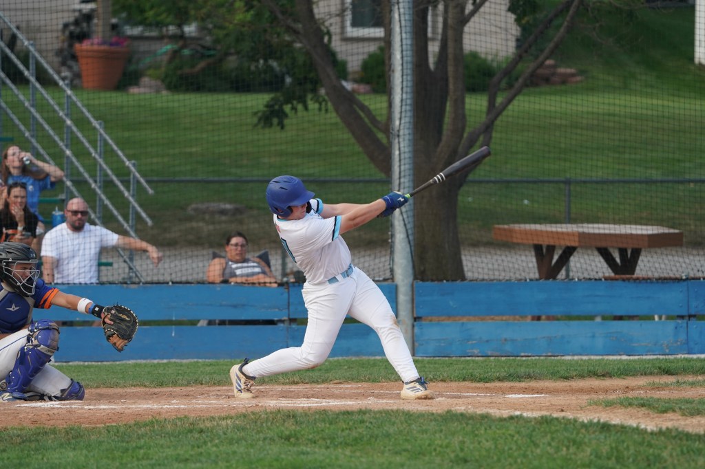 A young male baseball player in action, swinging a bat at a pitch. The catcher is crouched behind home plate, wearing protective gear. In the background, spectators watch the game, and a blue wooden bench is visible.