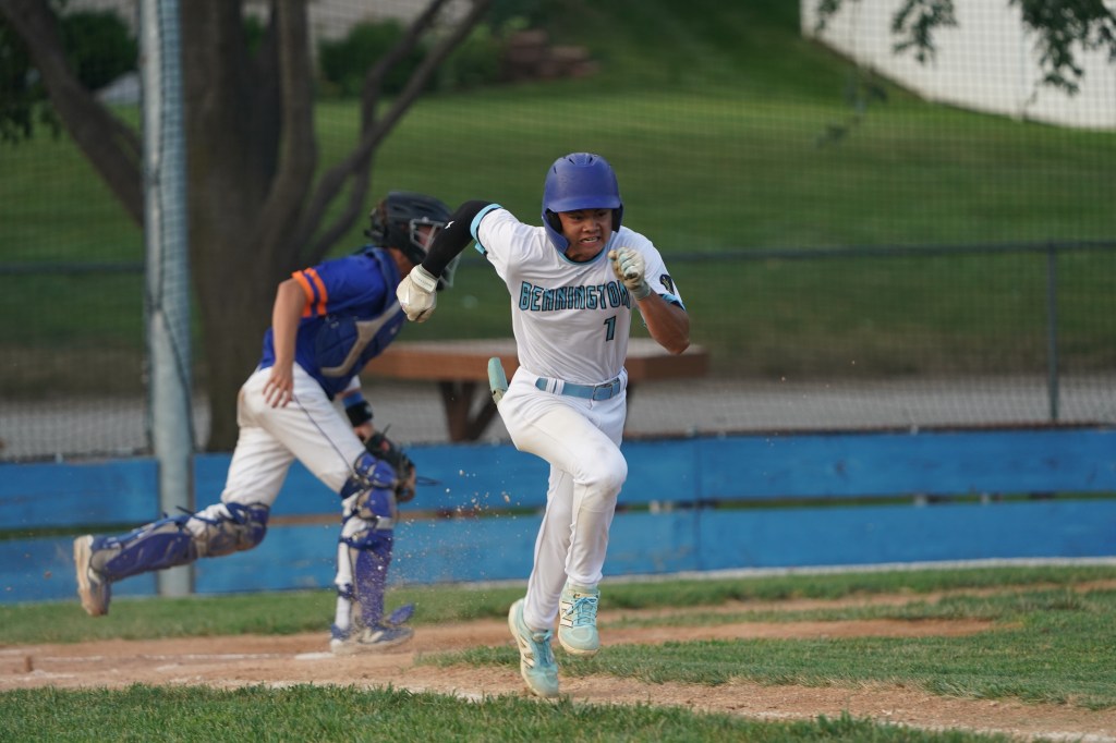 A baseball player wearing a white uniform with blue accents is sprinting toward home plate, while a catcher in a blue and orange uniform is positioned behind the plate.