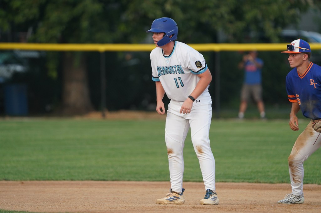 A young baseball player in a white uniform with the number 11 stands near second base on a baseball field. He has a blue helmet and is focused on the game while a teammate in an orange uniform stands nearby.