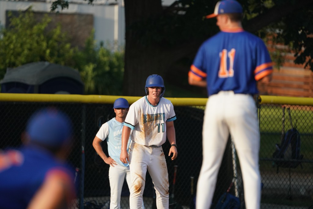 A baseball player in a white uniform with a blue helmet stands on base, looking toward the pitcher, while another player in a blue uniform with orange accents prepares to pitch.