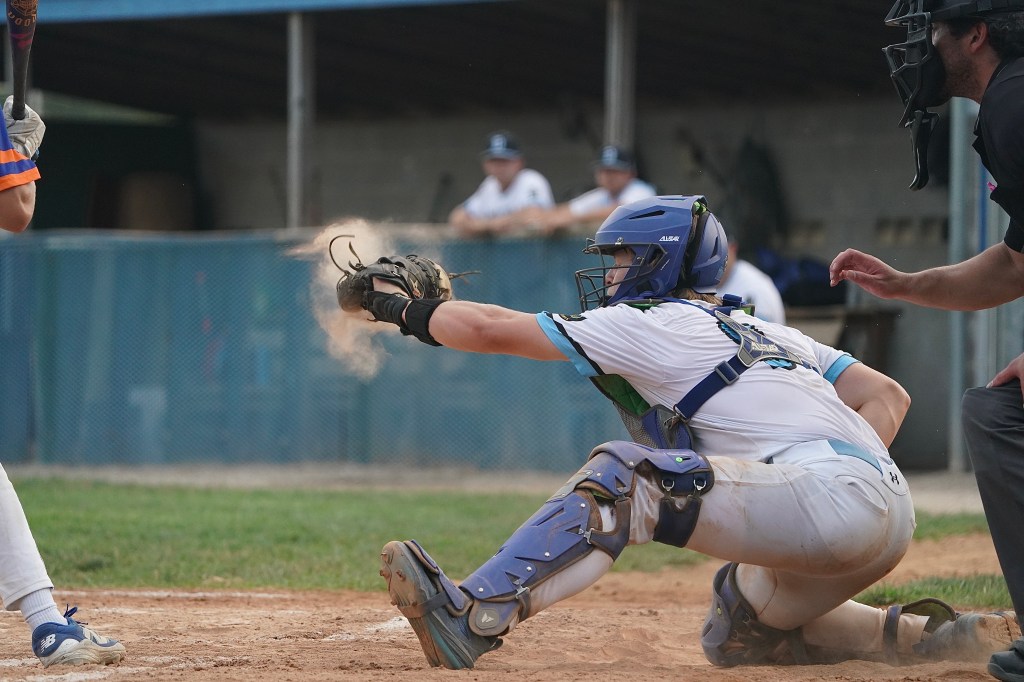 A youth baseball catcher crouches low, reaching for a pitch while dust kicks up around him. In the background, players watch from the dugout.