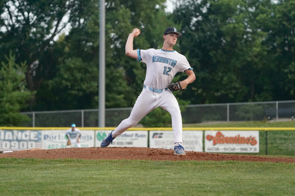 A baseball pitcher in a white uniform with blue accents, wearing a cap, is in the windup position on the pitcher's mound. The background shows a baseball field with trees and fencing.