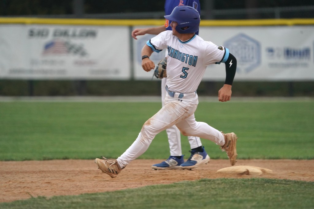 A baseball player in a white jersey with blue accents and the number 5 is sprinting towards first base on a dirt infield, with a teammate standing nearby.