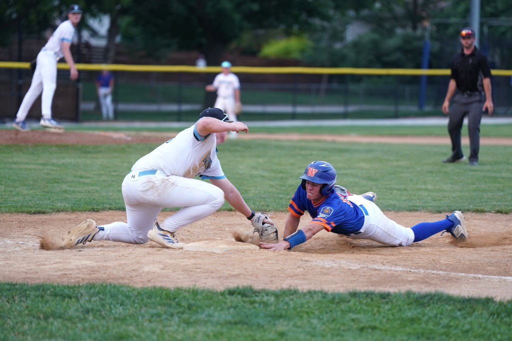 A baseball player sliding into home plate while another player attempts to tag him out. The scene captures the intensity of the game with players in uniforms and a grassy field.