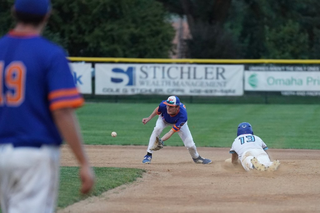 A baseball player in a blue and orange uniform attempts to field a ground ball while another player in white slides into base.