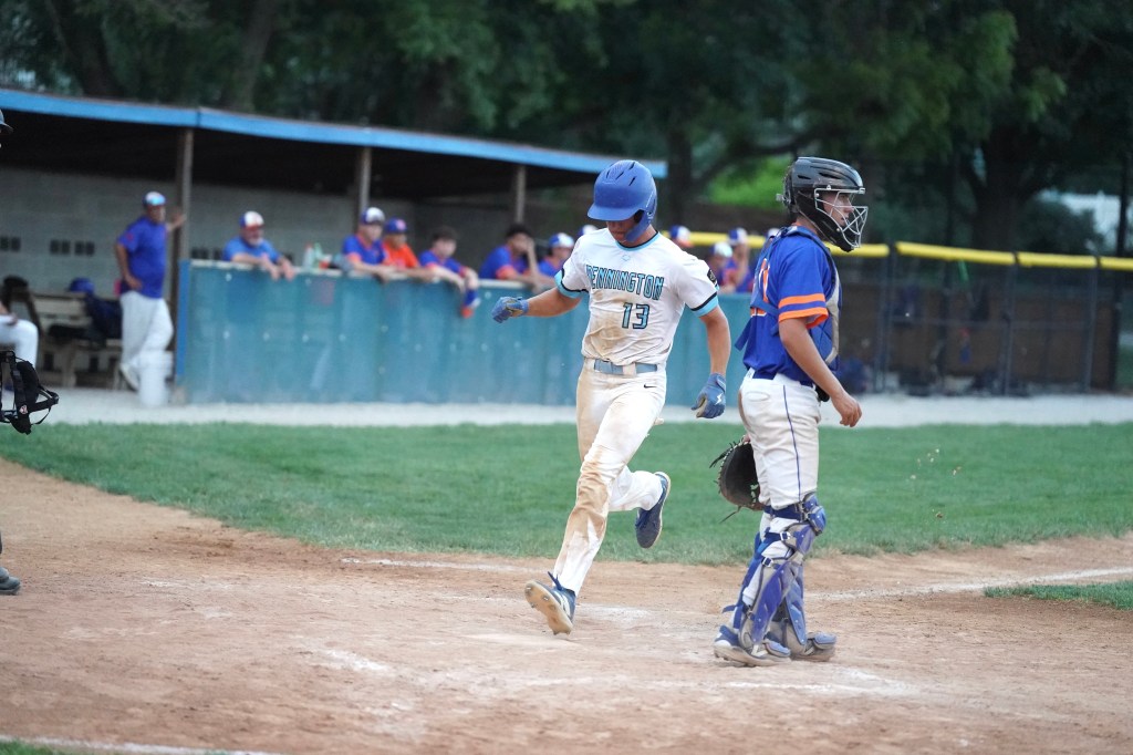 A baseball player in a blue uniform and helmet crosses home plate, celebrating after scoring a run, while a catcher in a blue jersey watches.