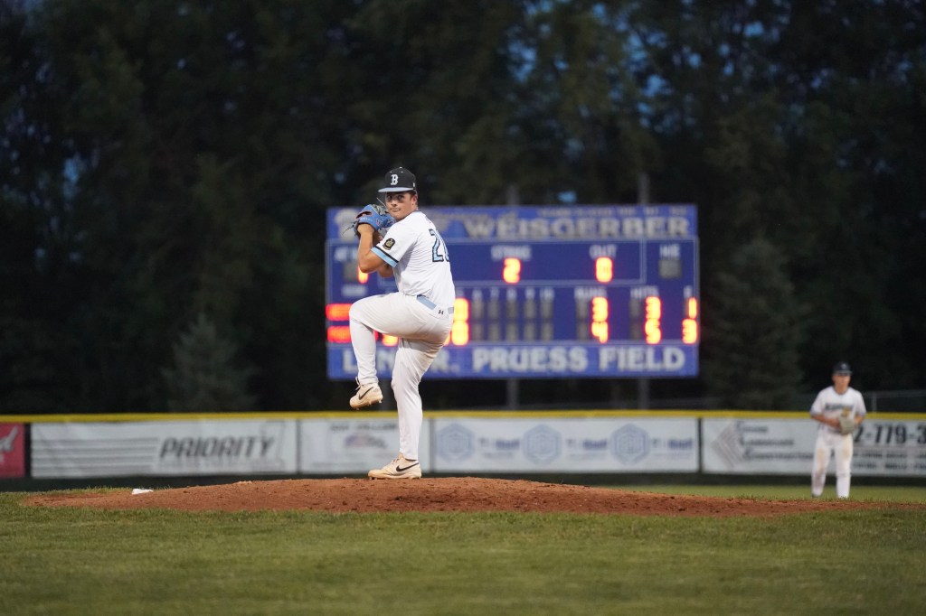 A baseball pitcher on the mound preparing to throw, with a scoreboard in the background displaying game statistics.