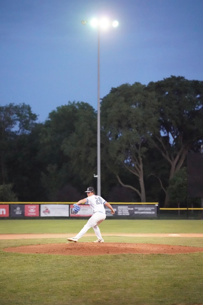 A baseball pitcher in a white uniform is winding up to throw on a dimly lit field during twilight.