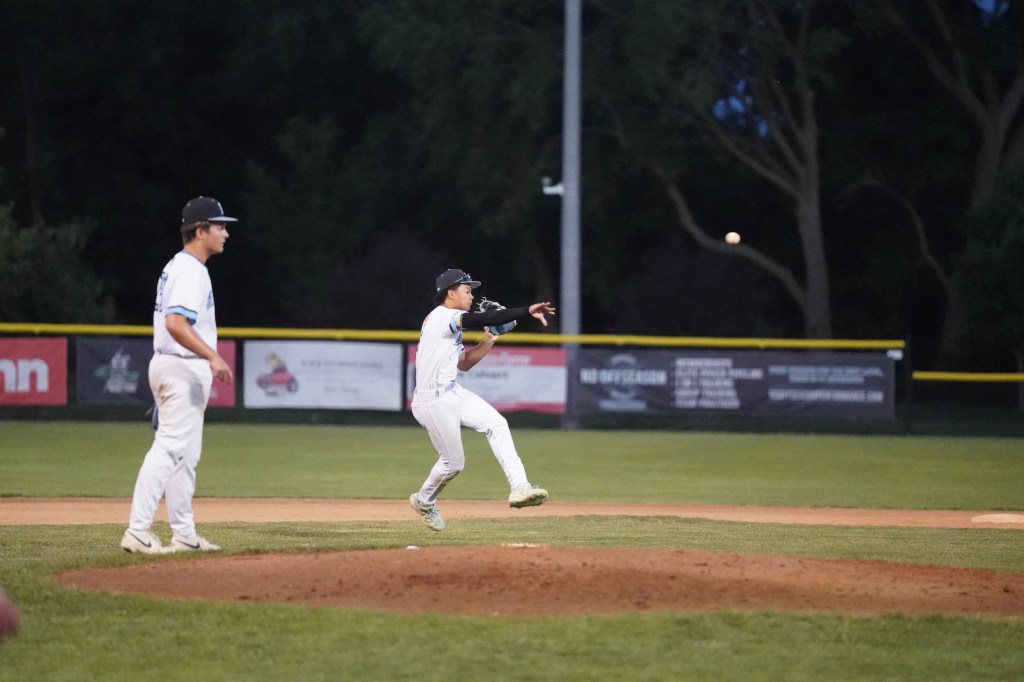 A young baseball player in a white uniform makes a catch while another player watches on the field during a night game.