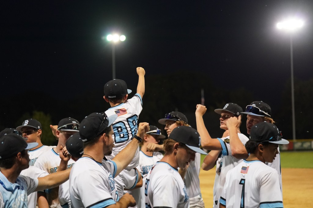 A youth baseball team celebrating with raised fists under stadium lights at night, while one player is lifted onto another's shoulders.