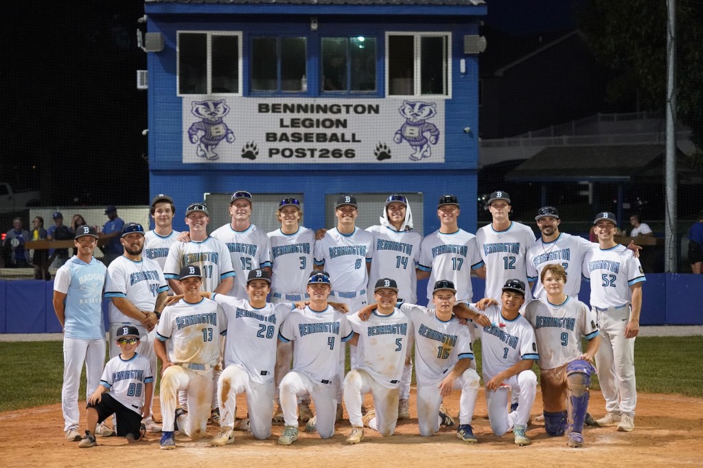 A group of baseball players posed together on the field in front of a blue press box featuring the Bennington Legion Baseball logo. The players are wearing white uniforms with light blue accents, displaying team pride.