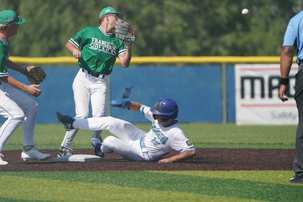A baseball player sliding into second base while two fielders in green uniforms prepare to make a play. The scene displays action on a baseball diamond with a blurred background of a blue sky and fencing.