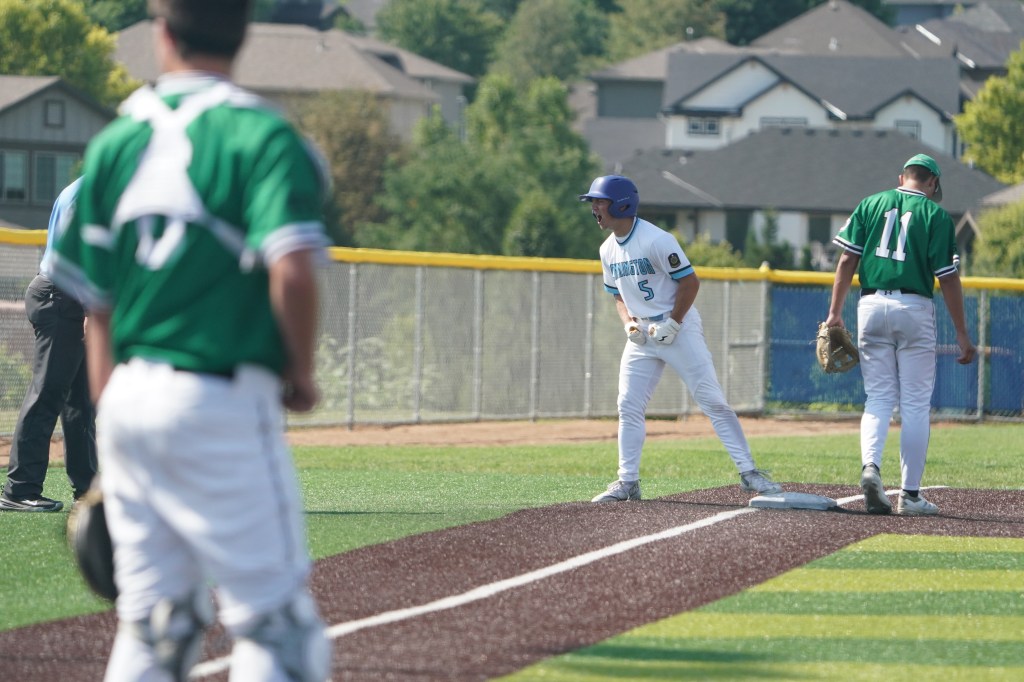 A baseball player in a blue jersey celebrating at first base, while two players in green jerseys observe from the field, with houses in the background.
