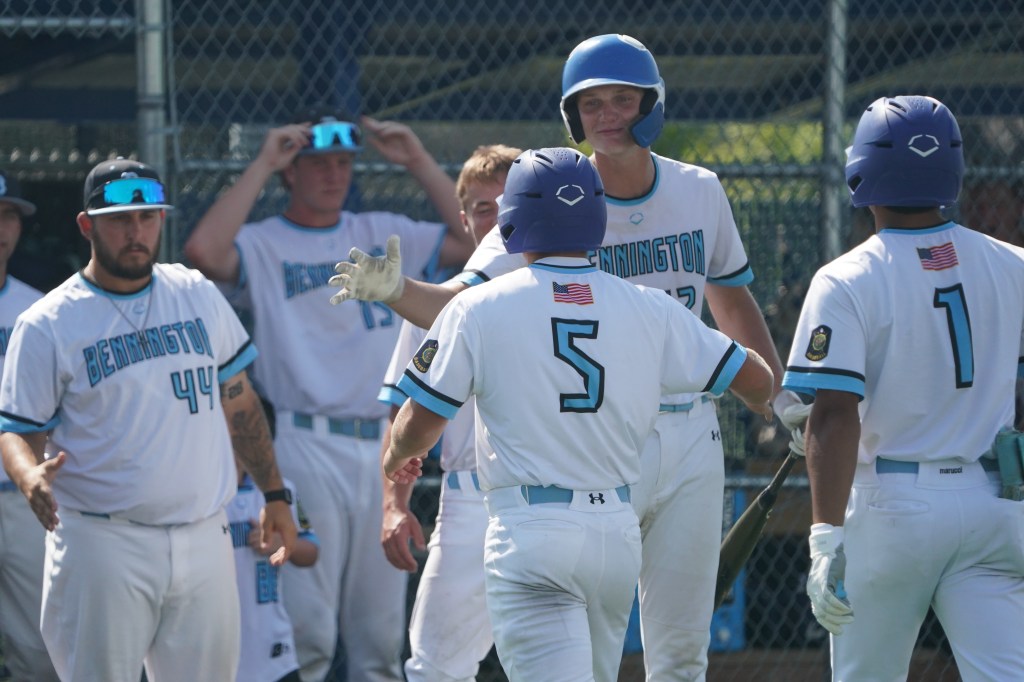 A group of baseball players from Bennington celebrate a play, with one player wearing jersey number 5 receiving congratulations from teammates in a sunny outdoor setting.