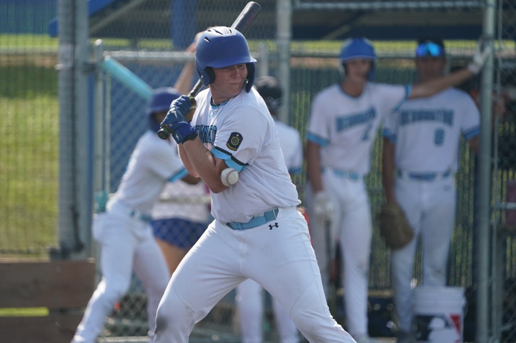 A baseball player in a white uniform with blue accents prepares to bat, with teammates visible in the dugout in the background.