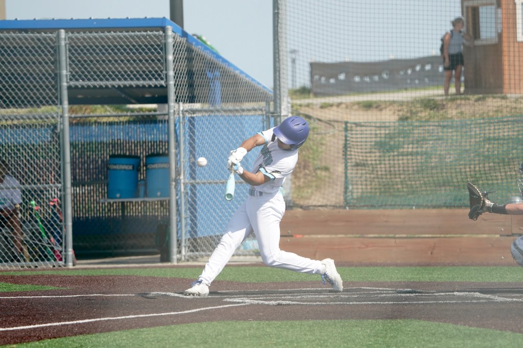 A baseball player swings a bat to hit a baseball during a practice or game. The player is dressed in a light blue jersey and white pants, wearing a helmet. The background includes fencing and blue containers.
