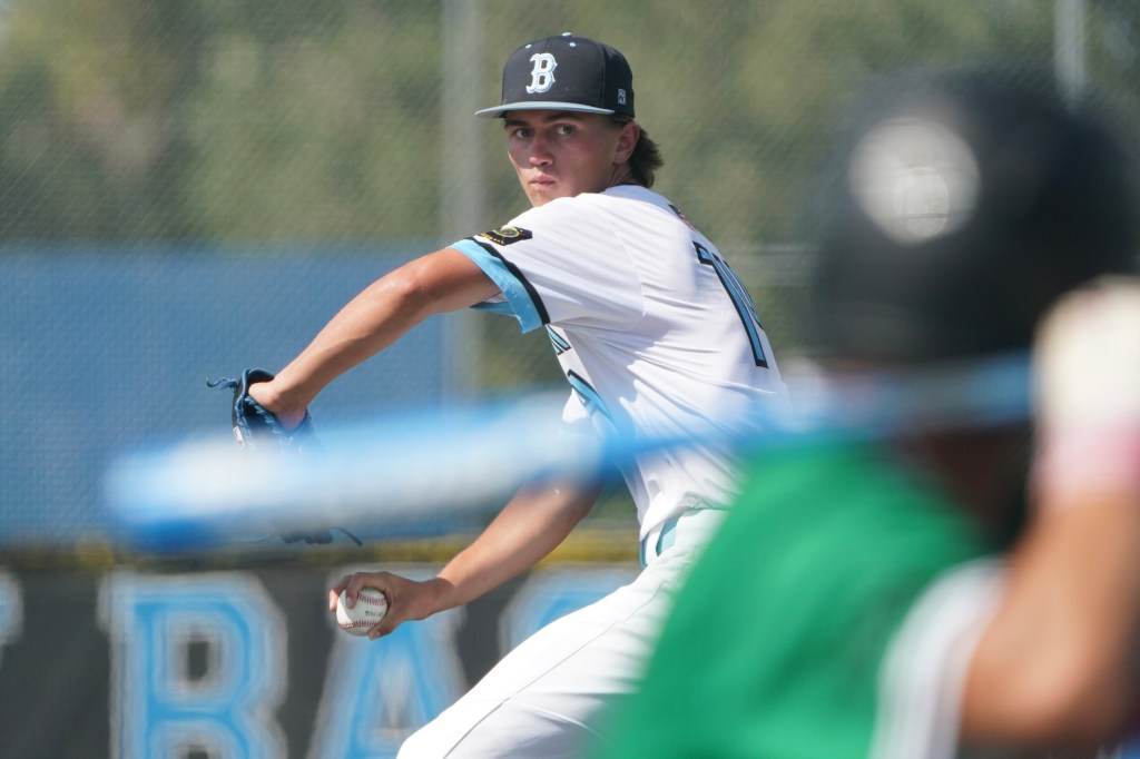 A baseball pitcher in a white uniform prepares to throw a pitch, with a blurred foreground player in a green jersey and a baseball in his hand.