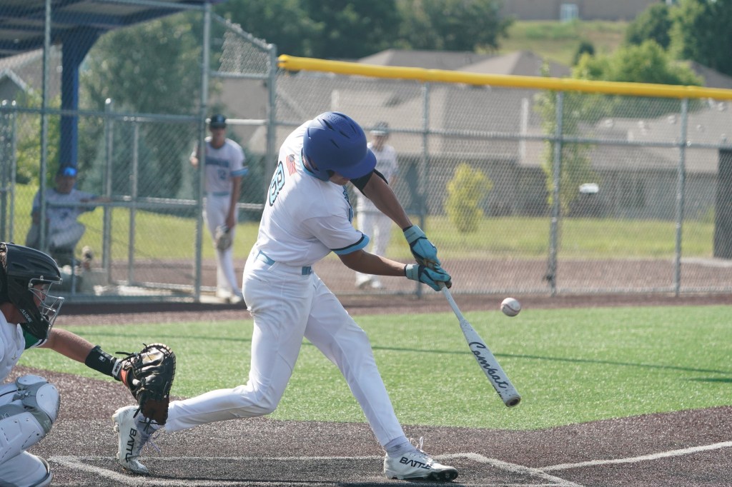 A young baseball player swings a bat to hit a baseball during a game, with players in the background and a sunny green field.