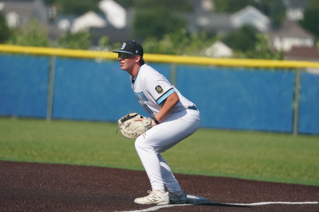 A baseball player in a white uniform is crouched in an athletic stance on the field, preparing to field a ball.