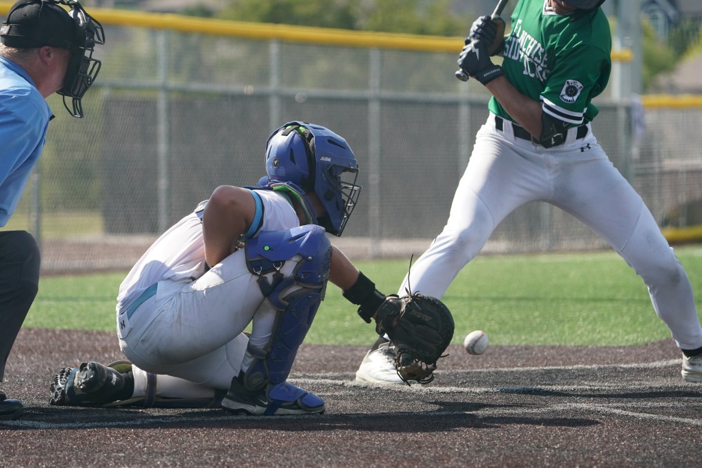 A baseball player crouches as a batter swings, with the ball heading towards the catcher in a game setting.
