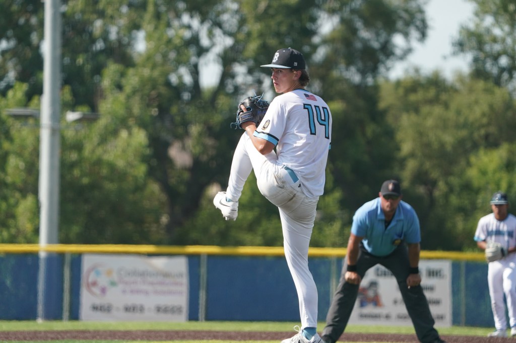 A baseball pitcher in a white uniform prepares to throw the ball, leg raised and arm back, with teammates and an umpire visible in the background.