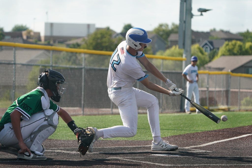 A baseball player wearing a white uniform swings a bat at a pitched ball, while a catcher in a green and white uniform crouches behind home plate.