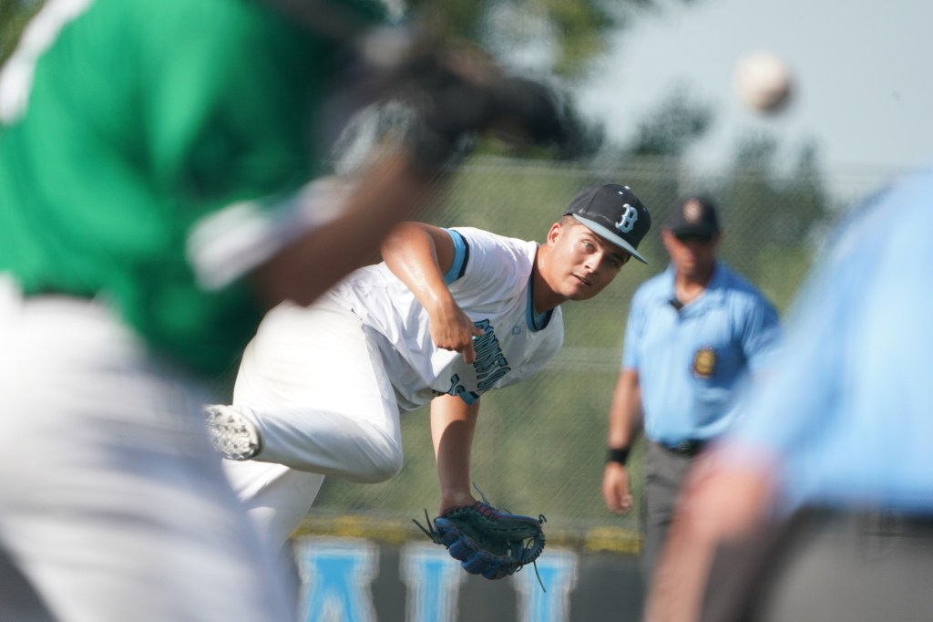 A young baseball pitcher in mid-throw, wearing a white jersey and black cap, with an opposing player in a green jersey visible in the foreground. An umpire watches in the background.