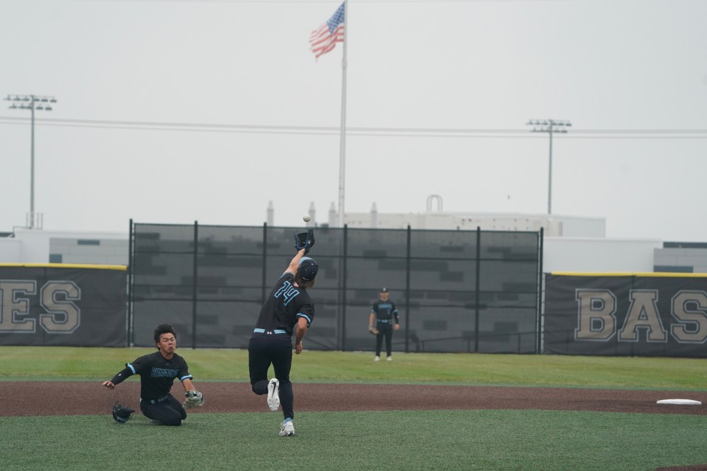 A baseball player in a black jersey is executing a throw while another player in a black jersey is on the ground, ready to catch the ball. An American flag is visible in the background.