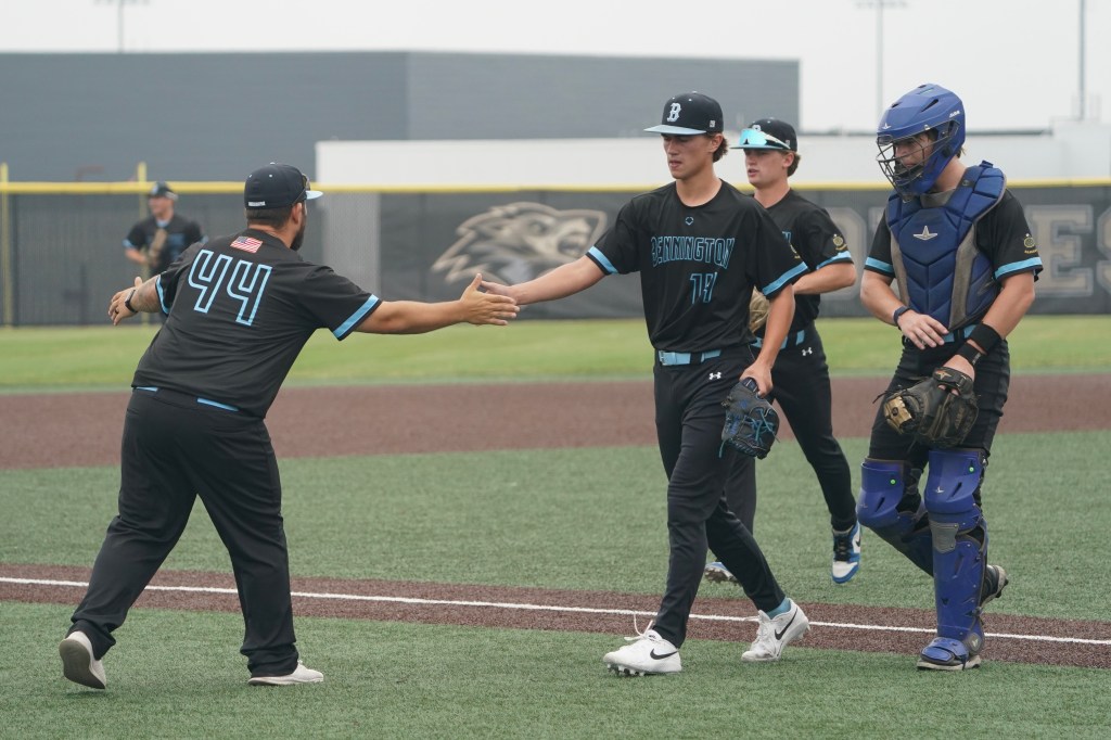 A baseball coach in a black shirt with the number 44 greets a player in a black uniform with the name 'Bennington' on it, as another player and a catcher in protective gear observe on the field.