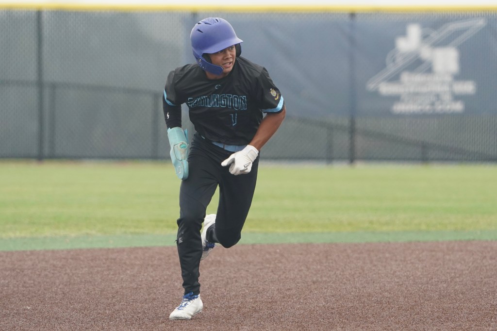 A young baseball player running on the field, wearing a dark uniform with blue accents and a purple helmet, focused and in motion.