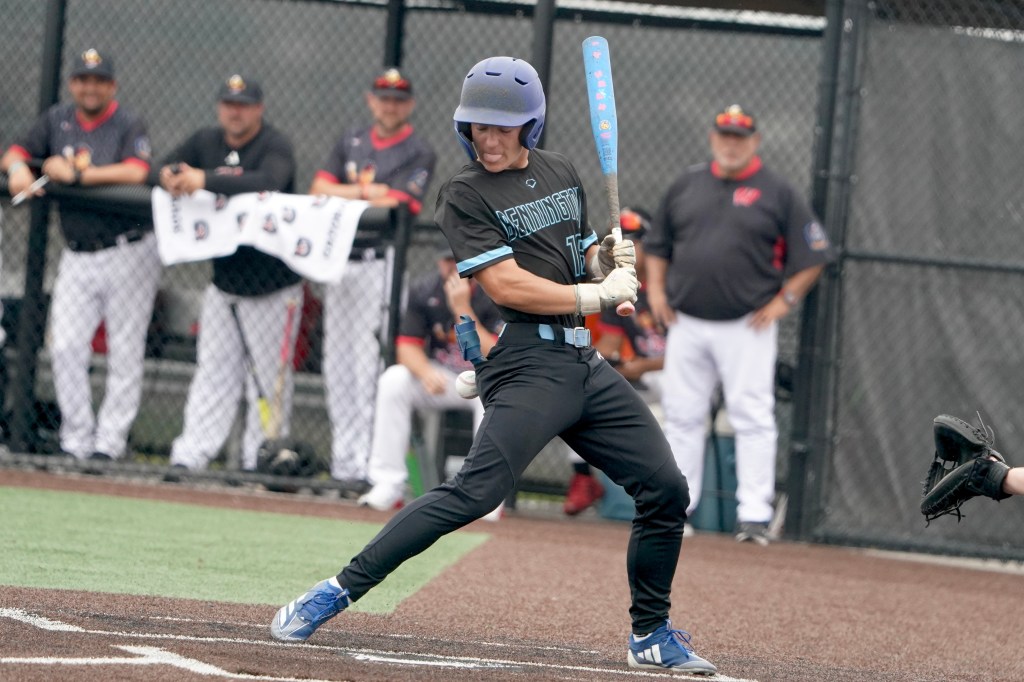 A baseball player in a black uniform swings a blue bat, preparing to hit the ball. Coaches and players are visible in the background.