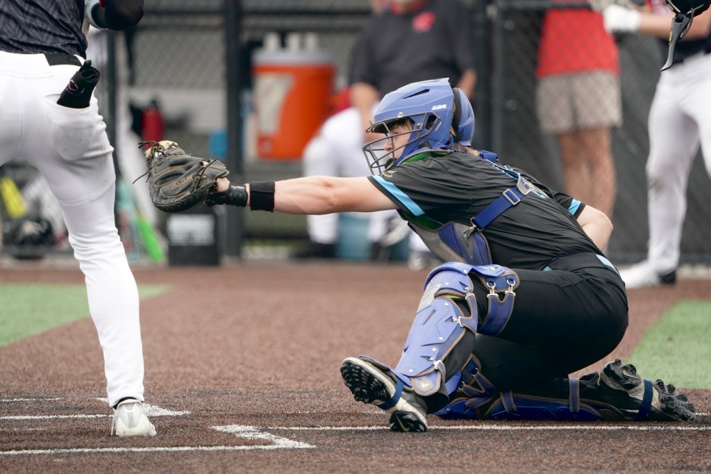 A baseball catcher in full gear crouches down, reaching out to catch a pitch while a batter stands in the background.