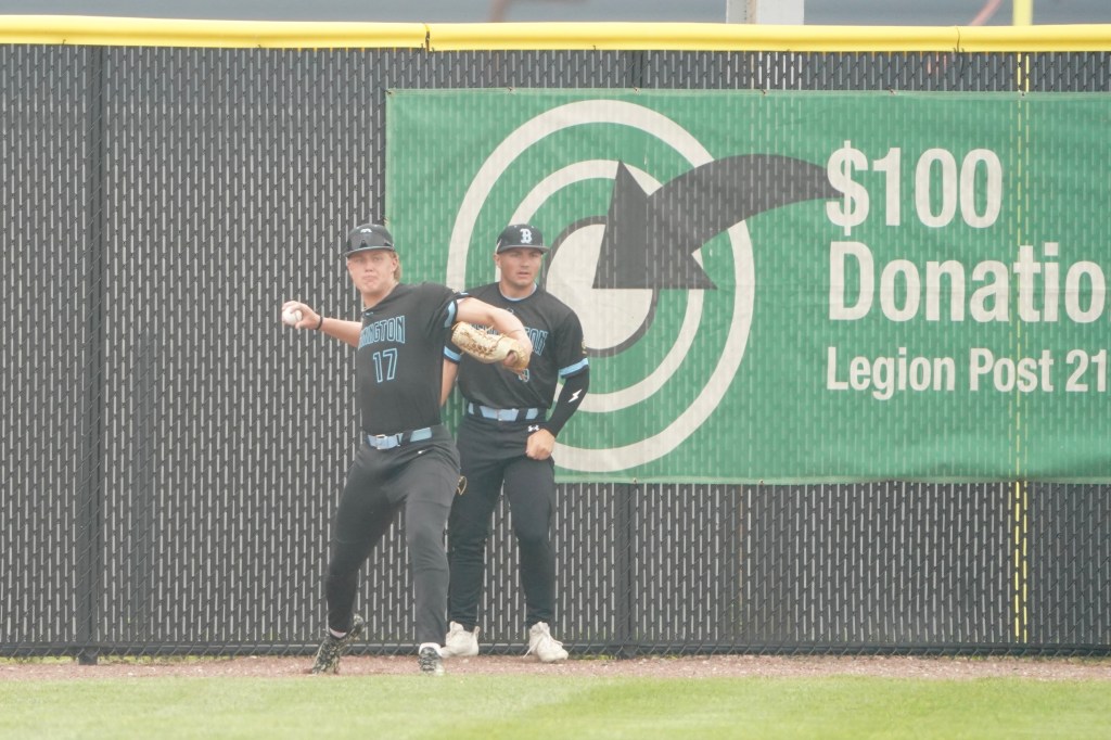 Two baseball players in black uniforms stand near a green donation sign on a sports field, with one player preparing to throw a baseball.