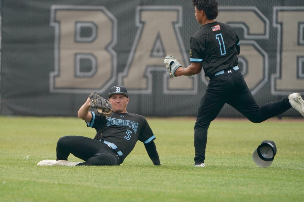 Two baseball players on the field; one player is sitting on the grass while catching a ball, and the other player is running towards him.