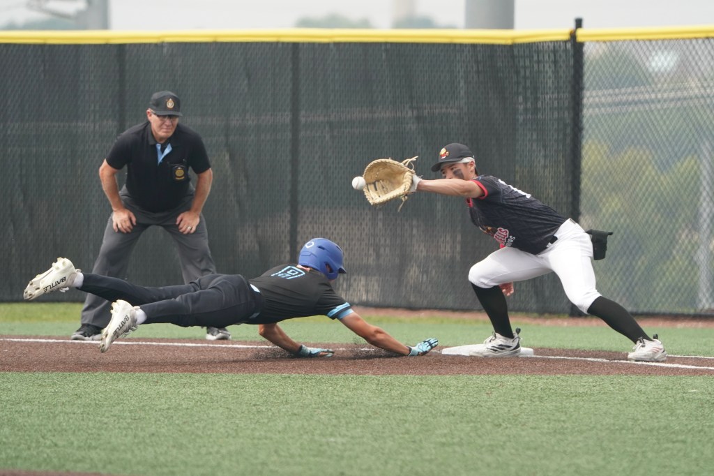A baseball player in a black uniform dives towards the base while another player in a white uniform reaches out with a glove to catch the ball. An umpire observes the play in the background.