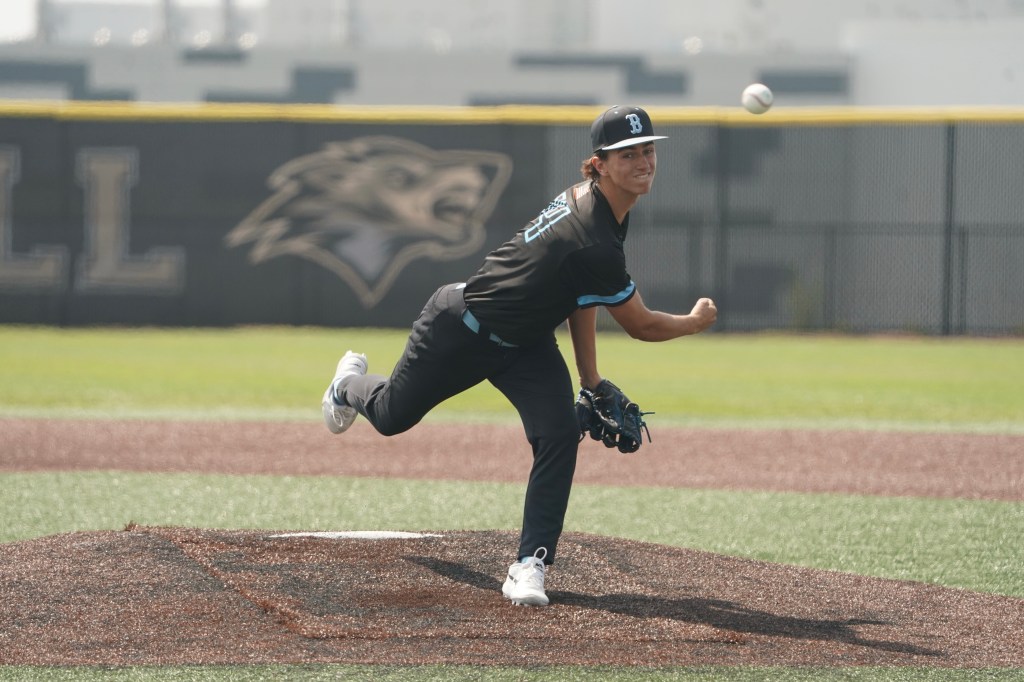 A young baseball player in a black uniform is pitching on a baseball field, with the ball in the air and a focused expression on his face.