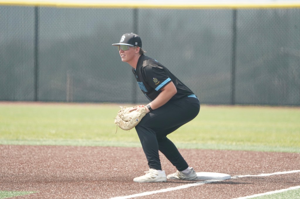 A young baseball player crouched at first base wearing a black jersey, black pants, and a cap, ready to react during a game.