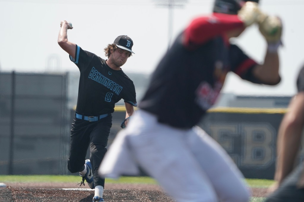 A baseball pitcher wearing a black jersey with 'Bennington' printed on it is in the windup position, preparing to throw a pitch. The focus is on the pitcher, while a batter in a red and black jersey is slightly blurred in the foreground.