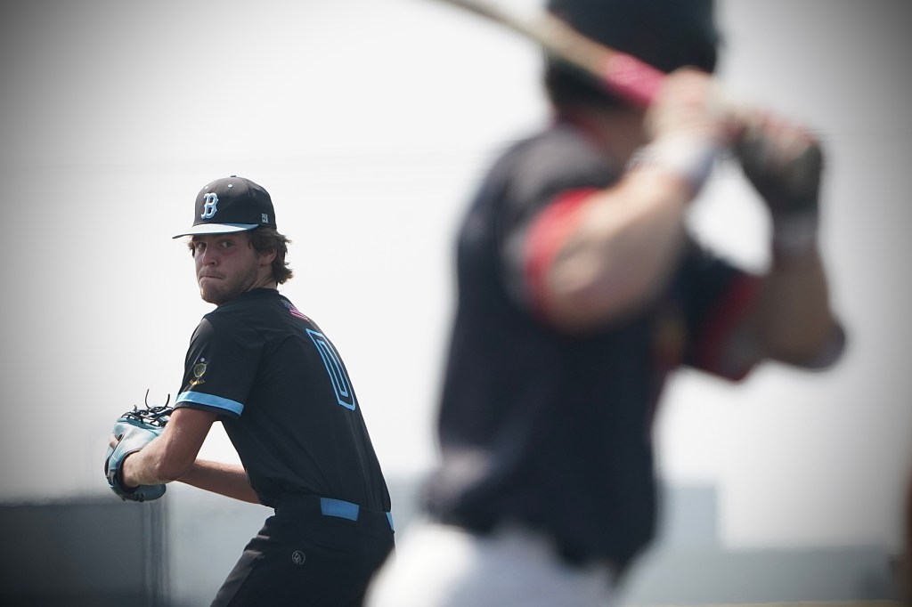 A baseball pitcher in a black uniform prepares to throw the ball, focusing intently. In the background, a batter stands poised to hit.