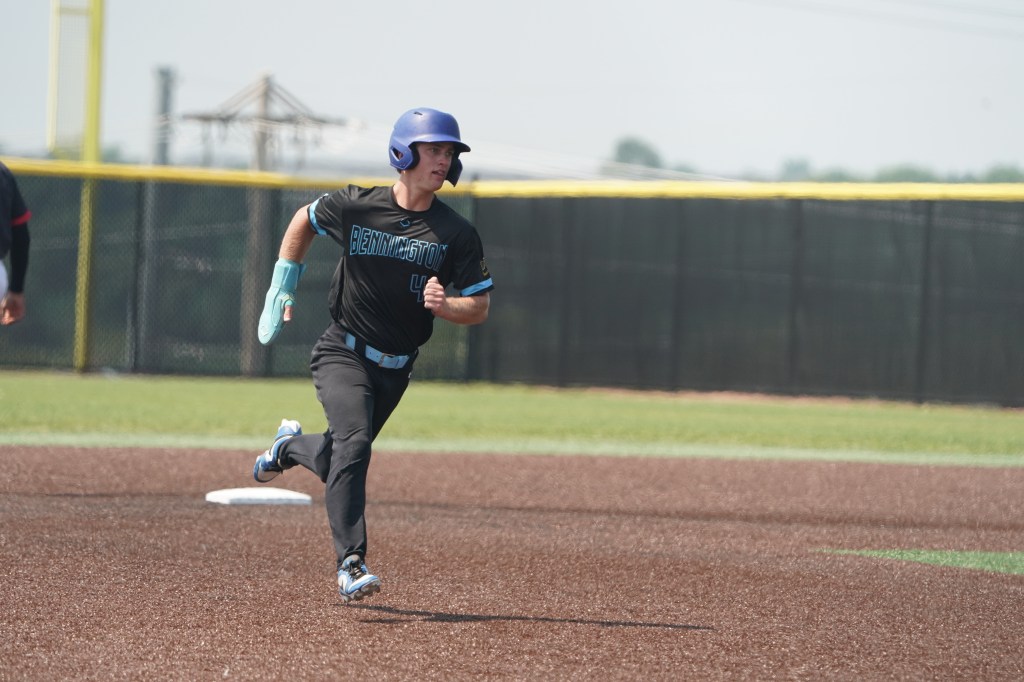 A baseball player wearing a black jersey with 'BENNINGTON' printed on it runs on the field, approaching first base, with a focused expression.