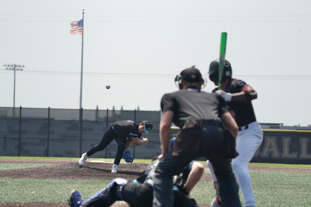 A baseball pitcher in a black uniform throws a ball towards a batter, who is ready to hit. An umpire crouches behind the batter, with an American flag visible in the background.