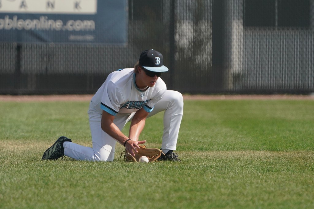 A young baseball player in a white uniform is kneeling on the grass, preparing to catch a ball.