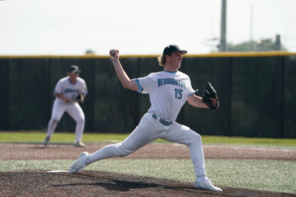 A young baseball pitcher in a white uniform with 'Bennington' on the chest is in the middle of a pitching motion on a baseball field. Another player stands in the background, ready for the play.