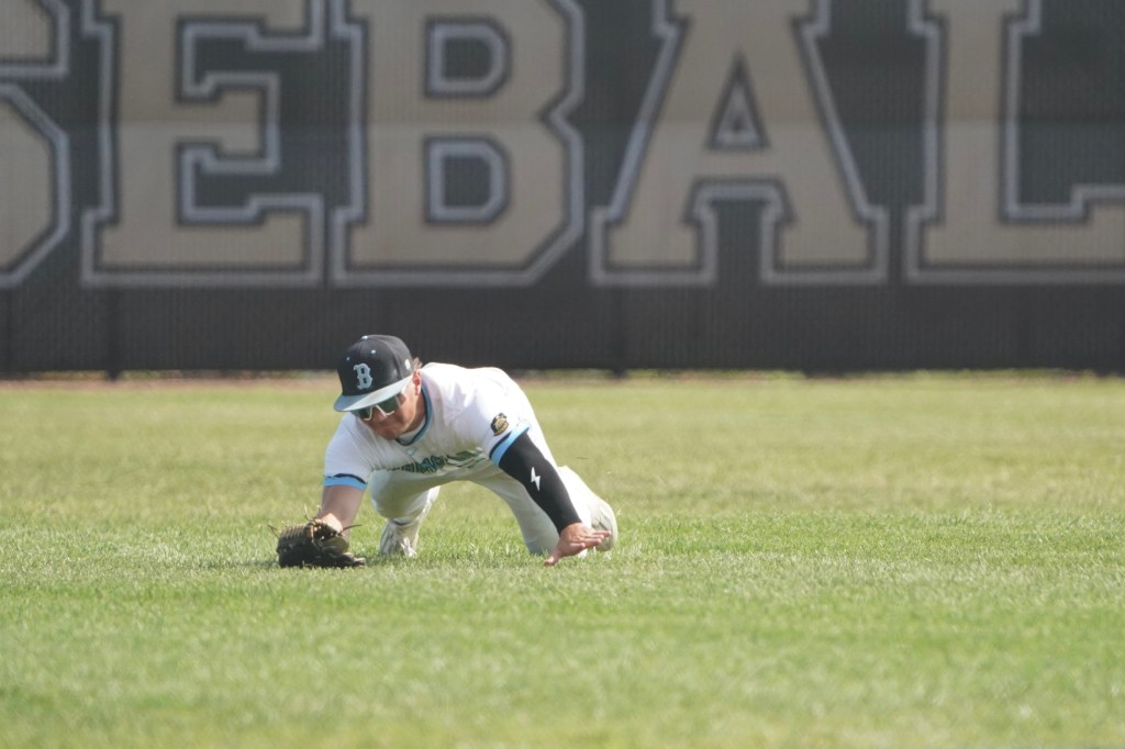 A baseball player diving to catch a ball on a grassy field, with a large 'BASEBALL' sign in the background.